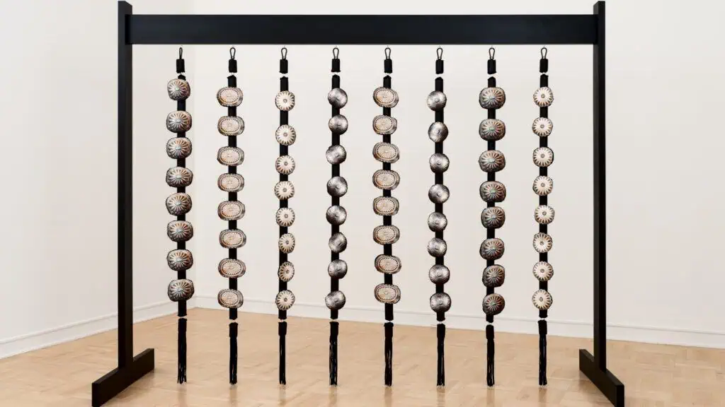 A black frame holds eight vertical rods, each threaded with round, patterned, ceramic disks, arranged in rows and displayed in a minimalist gallery space.