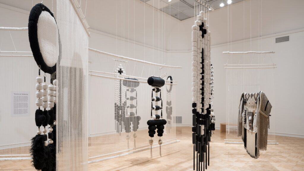 Art installation in a gallery featuring hanging textile sculptures and woven garments in black, white, and neutral tones, suspended from the ceiling with white string.
