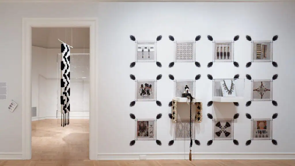 A gallery wall displays framed textile patterns, photographs, and objects, with an elongated black-and-white woven piece hanging in the adjacent room.