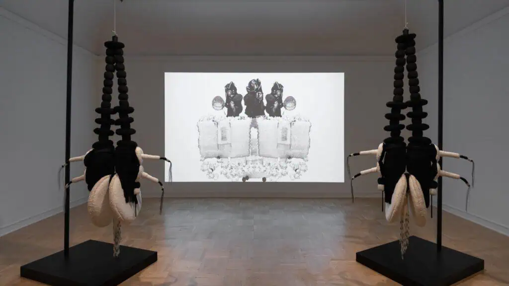 Two black sculptural installations with white appendages stand in a gallery space in front of a projected monochrome artwork on the back wall.