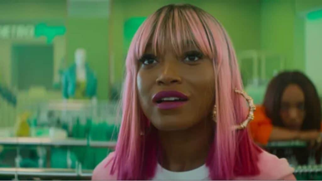 A woman with straight pink hair and large hoop earrings stands indoors, looking slightly upward; another person is blurred in the background.