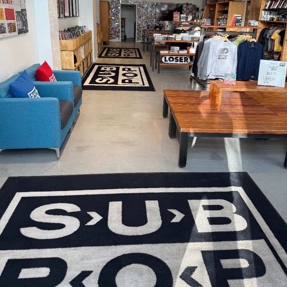 Retail store interior with SUB POP logo rugs, merchandise on display, a blue couch with pillows, and a wooden table in the center. Clothing and other items are arranged on shelves and racks.