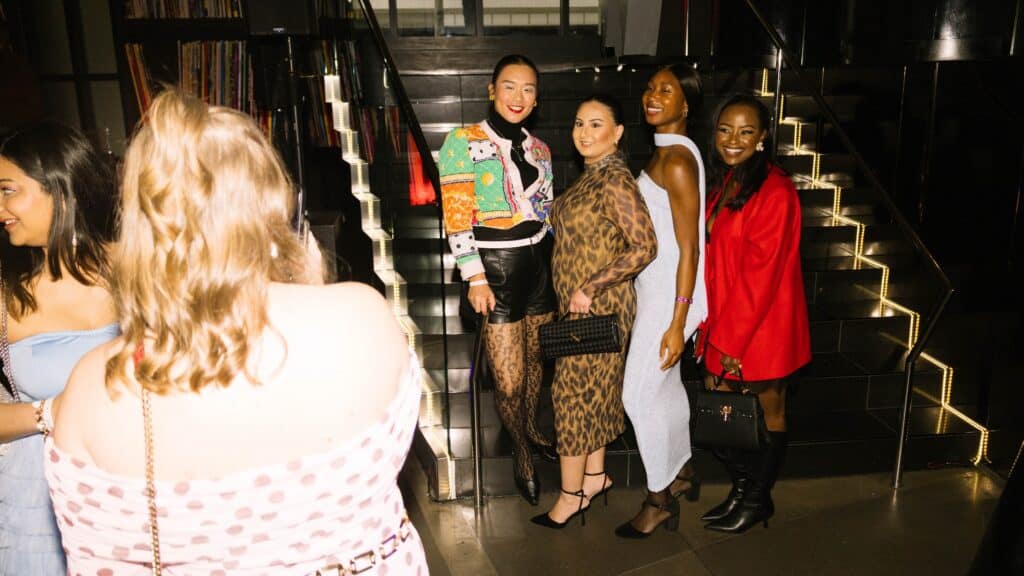 Four women pose for a photo on a lit staircase at SIFF, while another woman in a polka dot dress stands in the foreground, her back to the camera as if capturing the scene in her rearview mirror.