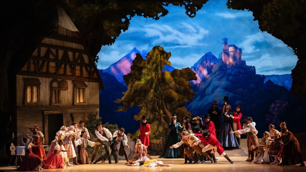 A large group of ballet dancers in elaborate costumes perform a dramatic scene on stage, capturing ballet’s saddest story against a painted village and mountain backdrop.