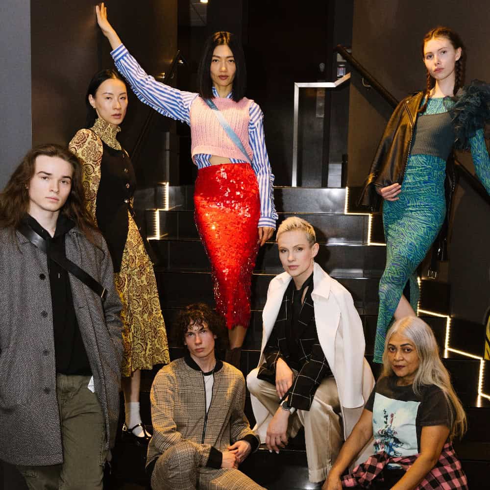 Six people pose on a staircase, wearing a variety of fashionable and eclectic clothing styles, with neutral to serious expressions.