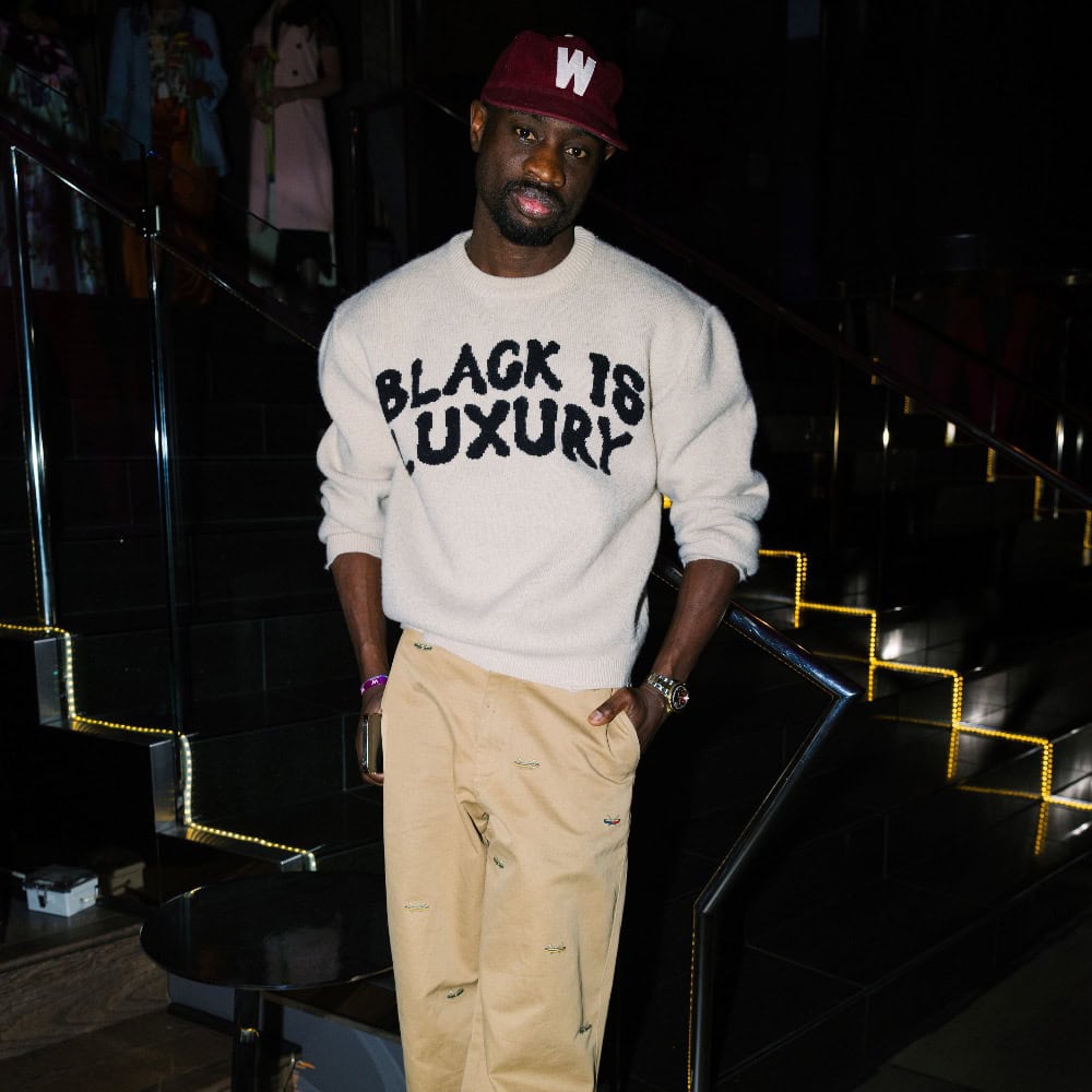 A man stands on a staircase wearing a maroon cap, a white sweater with "BLACK IS LUXURY" text, and tan pants—perfect inspiration for new outfit ideas to refresh your closet.