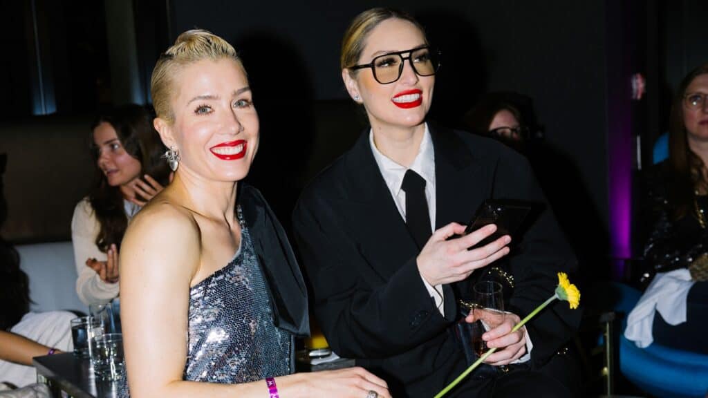 Two women with bright red lipstick sit together at a SIFF event; one wears a sparkly dress, the other a black suit with oversized glasses, holding a phone and a yellow flower—a striking pair reflecting style and family bonds.
