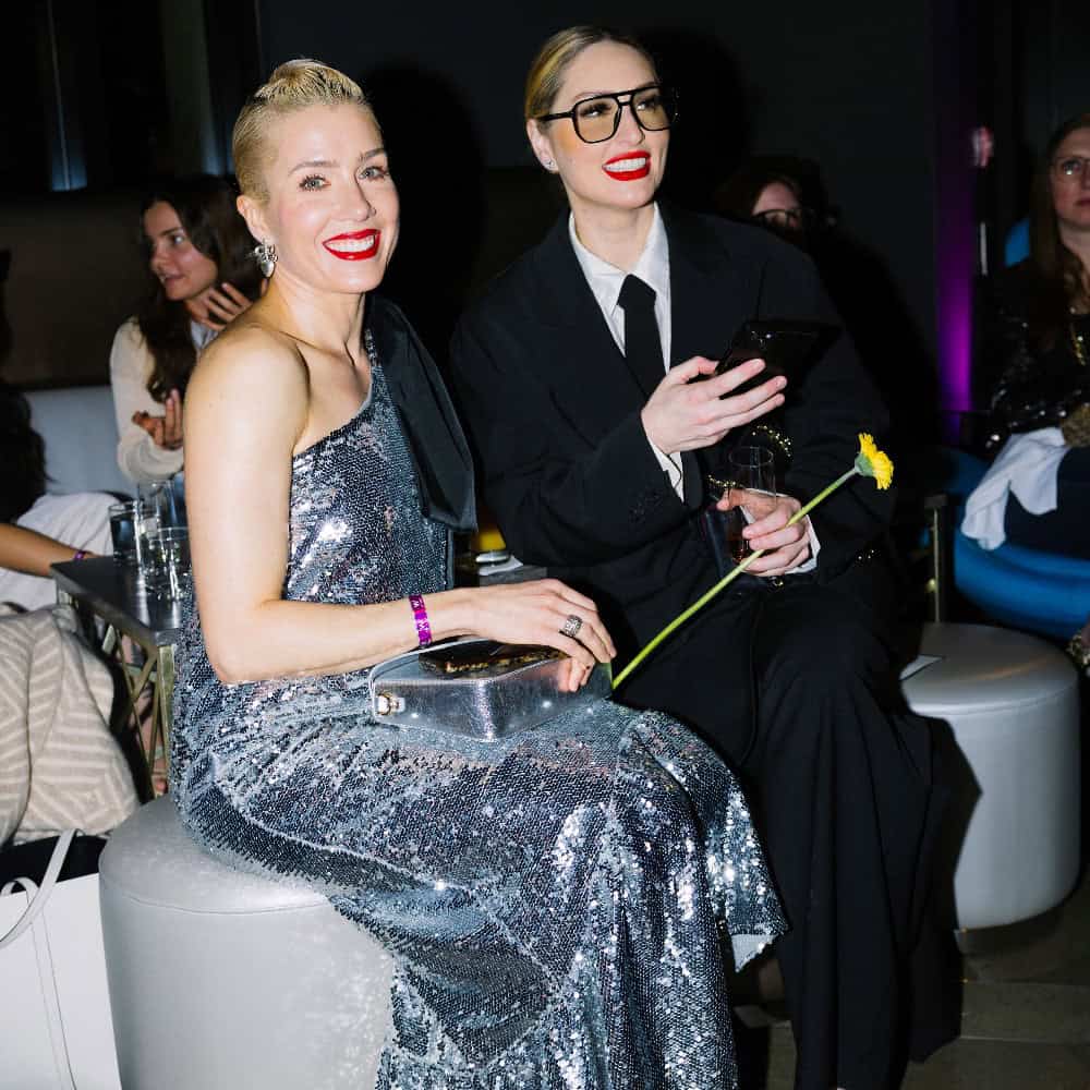 Two women sit side by side at an event; one wears a silver sequin dress and holds a clear clutch, the other wears a black suit and holds a yellow flower and a phone.