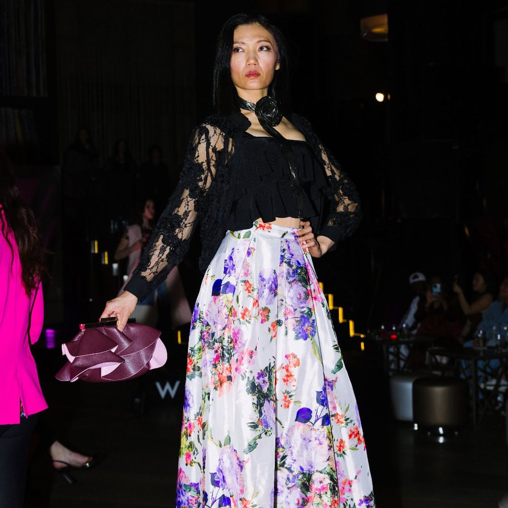 A woman stands on a runway wearing a black lace top and a long, floral skirt, holding a mauve hat in one hand. The background is dimly lit with seated spectators.