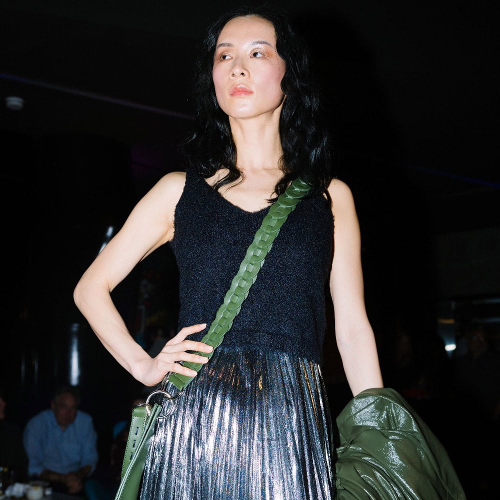 A person with long black hair wearing a sleeveless dark top, metallic silver pleated skirt, and green crossbody bag stands indoors holding a green jacket.