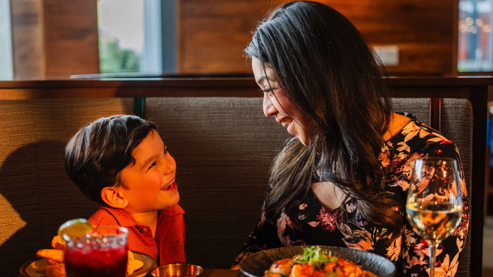 A woman and a young boy sit in a restaurant booth, smiling at each other with food and drinks on the table in front of them.