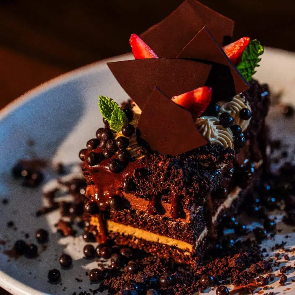 A slice of chocolate layer cake topped with strawberries, mint leaves, chocolate shards, and chocolate pearls on a white plate—perfect for a Mother’s Day brunch or your next Brunch Bonanza.