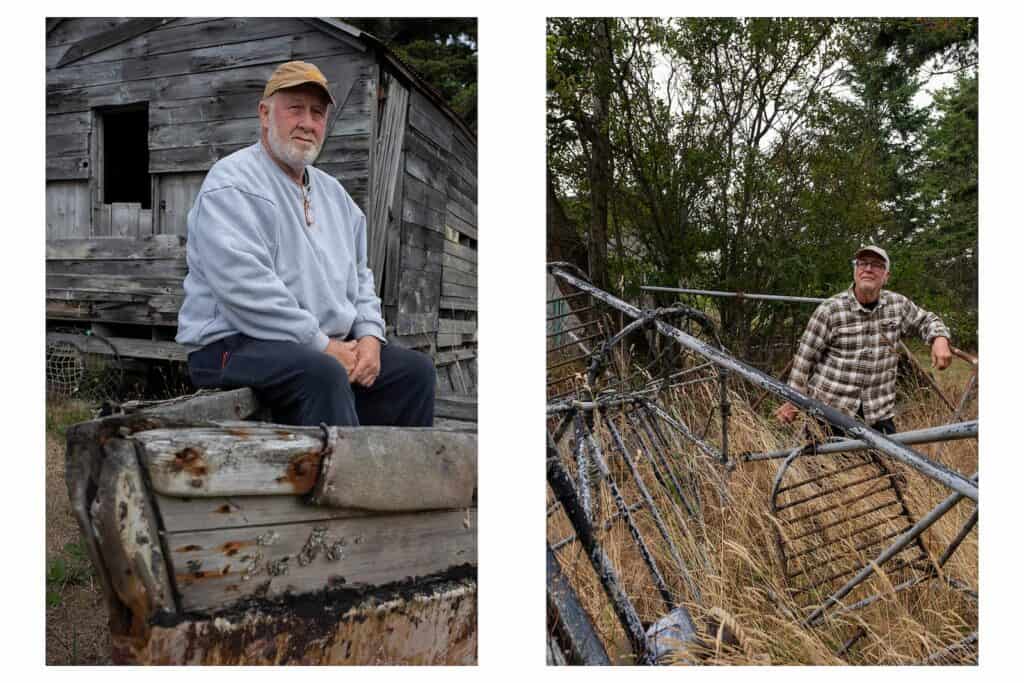 Two older men outdoors: one sitting on a weathered boat in front of a rundown wooden building, the other standing beside rusted metal debris in tall grass.