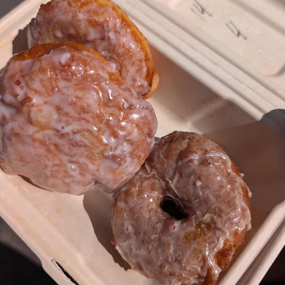 Three glazed donuts are placed inside a beige takeout container, with sunlight highlighting the shiny glaze on their surface.