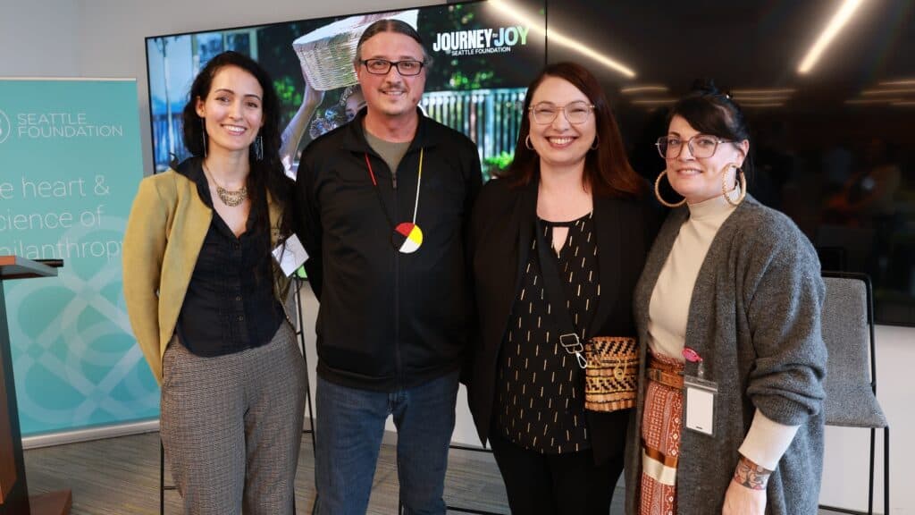 Four people standing side by side, smiling at the camera at an indoor event with a Seattle Foundation banner and TV screen in the background.