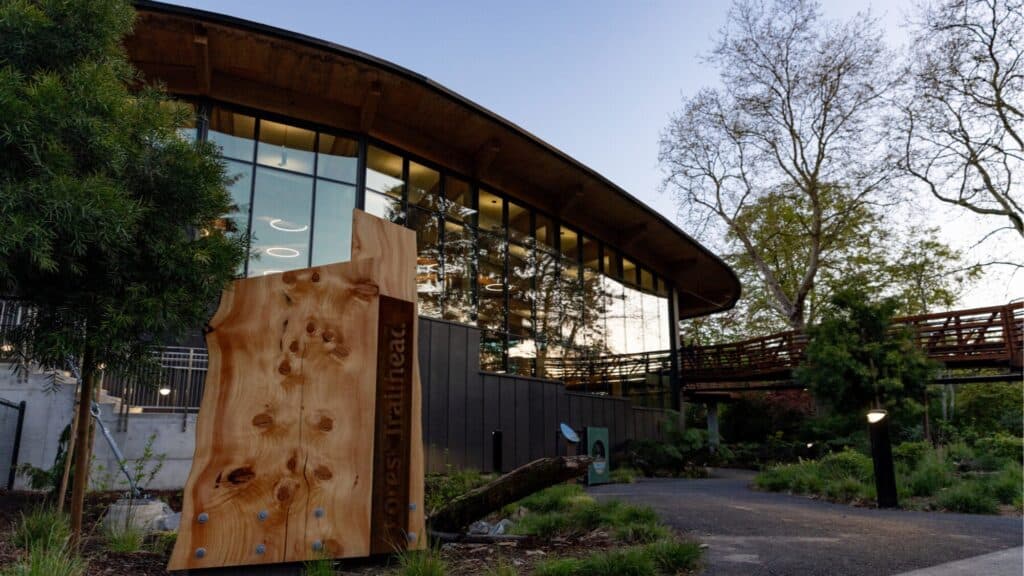 A modern building with large windows is surrounded by trees and greenery; a wooden sign labeled “Forest Trailhead” stands near a paved walkway at dusk, just steps away from the cozy charm of Waterfront Coffee.