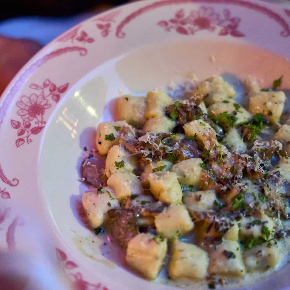 A plate of gnocchi topped with creamy sauce, mushrooms, grated cheese, and chopped herbs is served on a decorative white and pink dish—perfect for enjoying April food dishes in style.