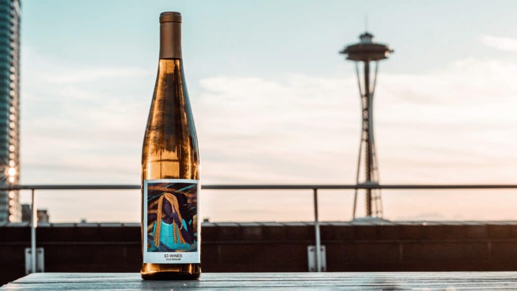 A bottle of home-grown wine label sits on a table outdoors, with the Space Needle and city skyline near sculpture park blurred in the background at sunset.