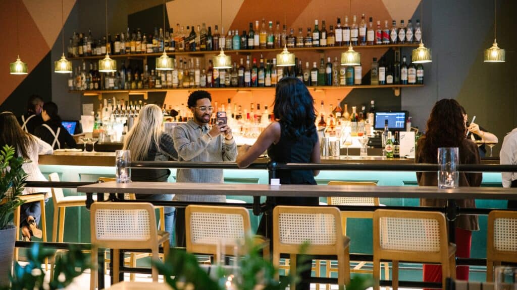 People are socializing at a modern bar, with bottles lined up on shelves behind the counter and pendant lights hanging overhead.