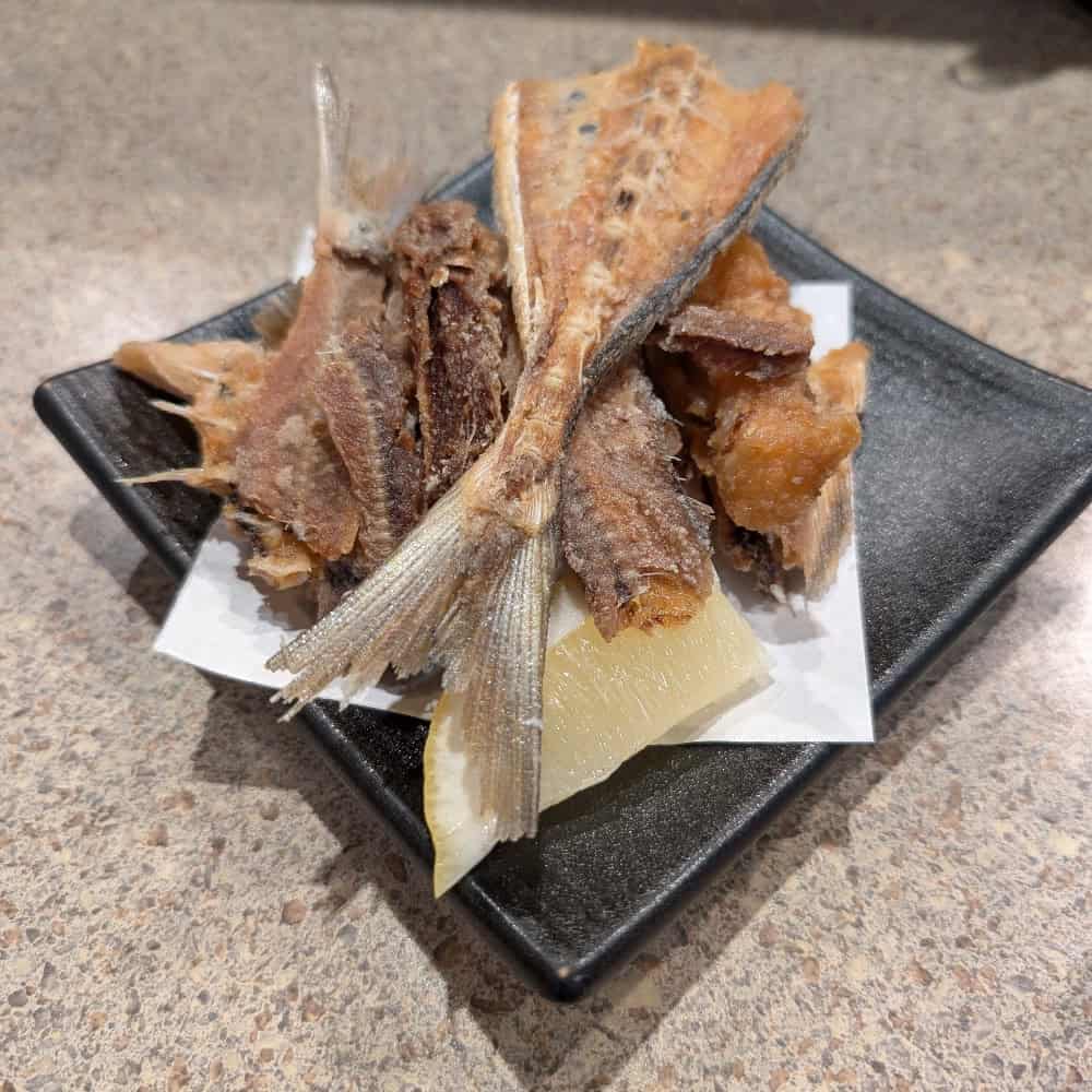 A black square plate with fried fish bones and pieces placed on a white paper, set on a speckled countertop.