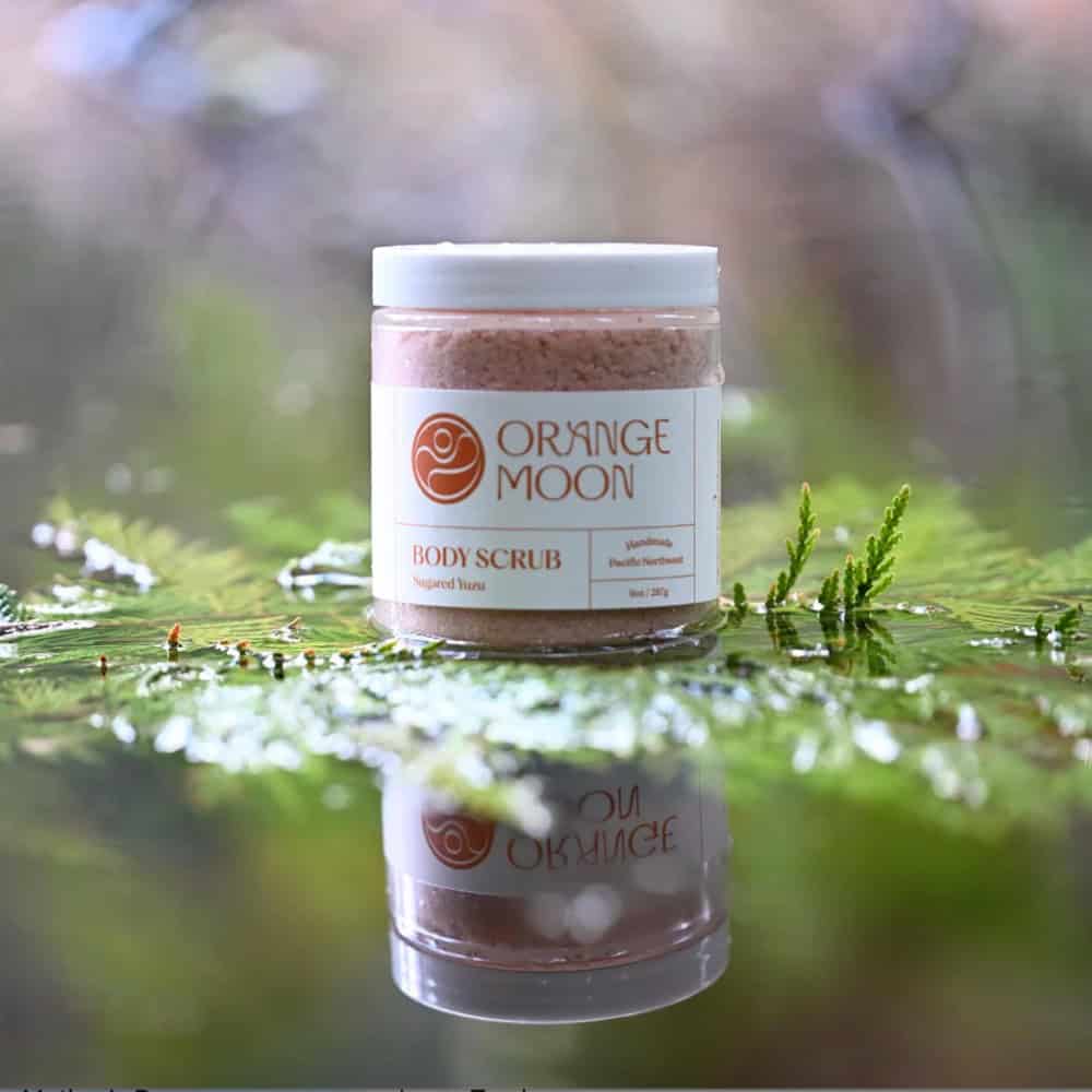 A jar of Orange Moon body scrub is placed on a reflective surface outdoors, surrounded by green moss and foliage.