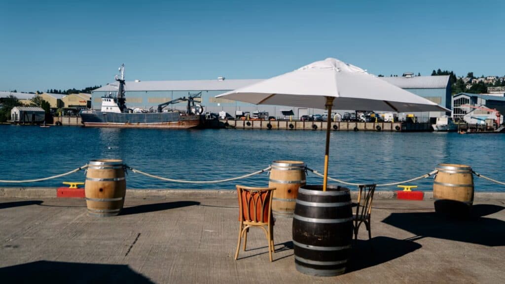 A waterfront dock with wooden barrels and chairs arranged under a white umbrella, hosting a relaxed rally as boats glide by and industrial buildings rise across the water—a scene reminiscent of Angela Dunleavy’s vibrant gatherings.