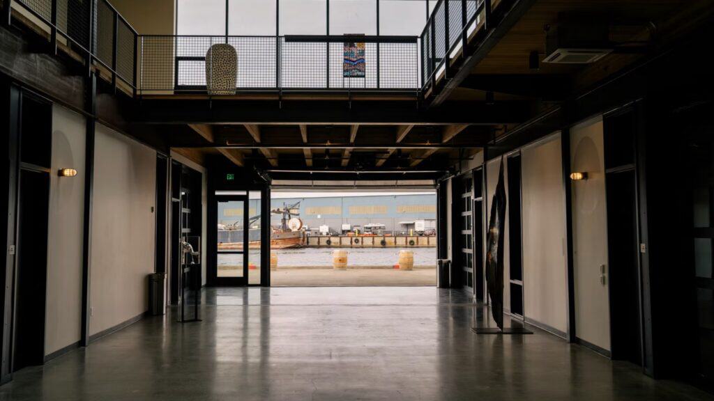 A spacious indoor hallway with polished floors leads to large glass doors, revealing a docked boat and industrial buildings outside—an ideal setting for an Angela Dunleavy Rally event.