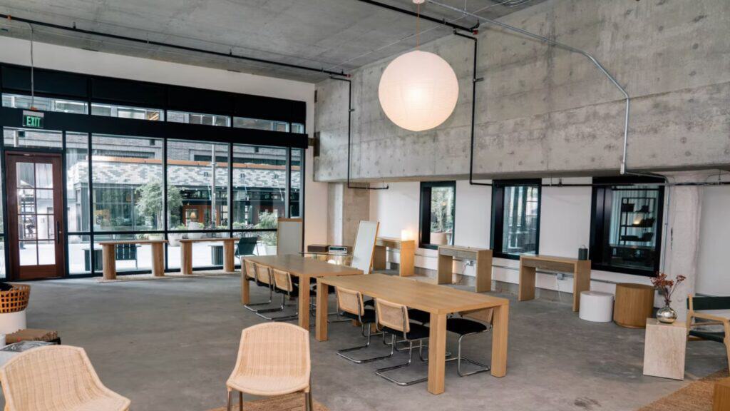 Modern open-plan office space with wooden tables, wicker and black chairs, large windows, concrete walls, and a round hanging light fixture.