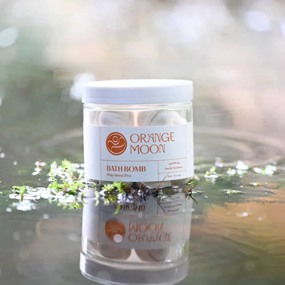 A clear jar labeled "Orange Moon Bath Bomb Pink Sweet Pea" sits on a reflective surface with small green plants around it—an ideal Mother’s Day gift to pamper someone special.