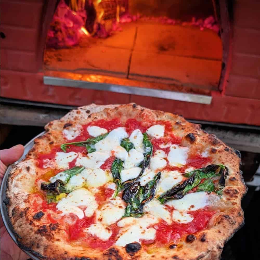A hand holds a freshly baked Margherita pizza with melted cheese, tomato sauce, and basil in front of a wood-fired oven.