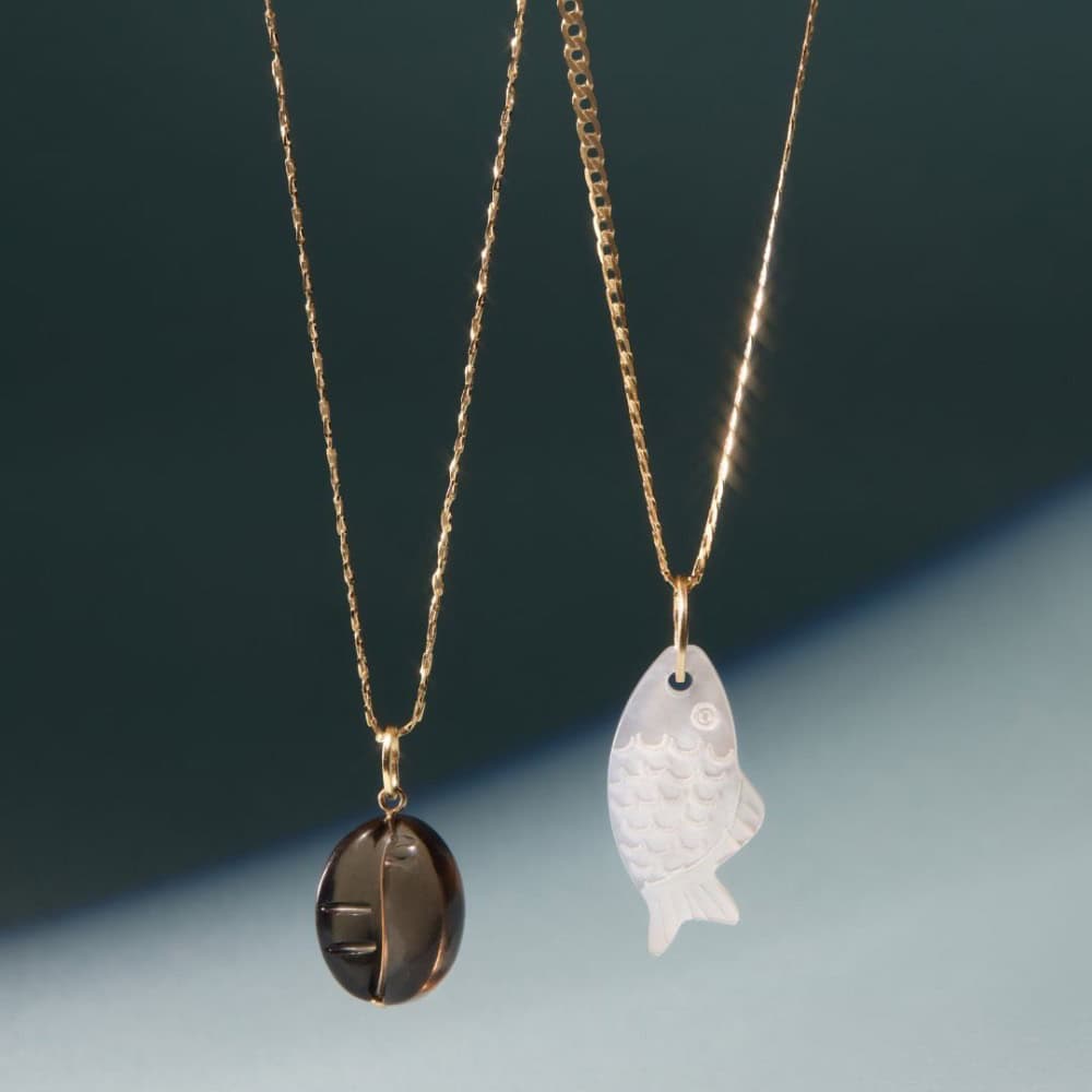 Two gold necklaces with pendants: one features a dark, round, carved stone; the other displays a translucent, carved fish. Both are shown against a soft, gradient background.