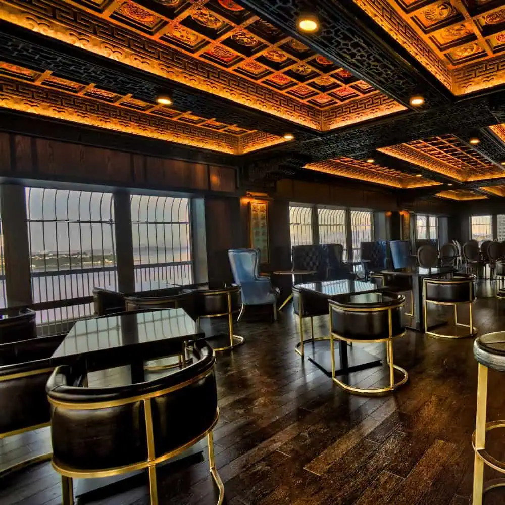 A luxurious lounge with dark wood floors, ornate ceiling, modern black and gold chairs, large windows, and river views.