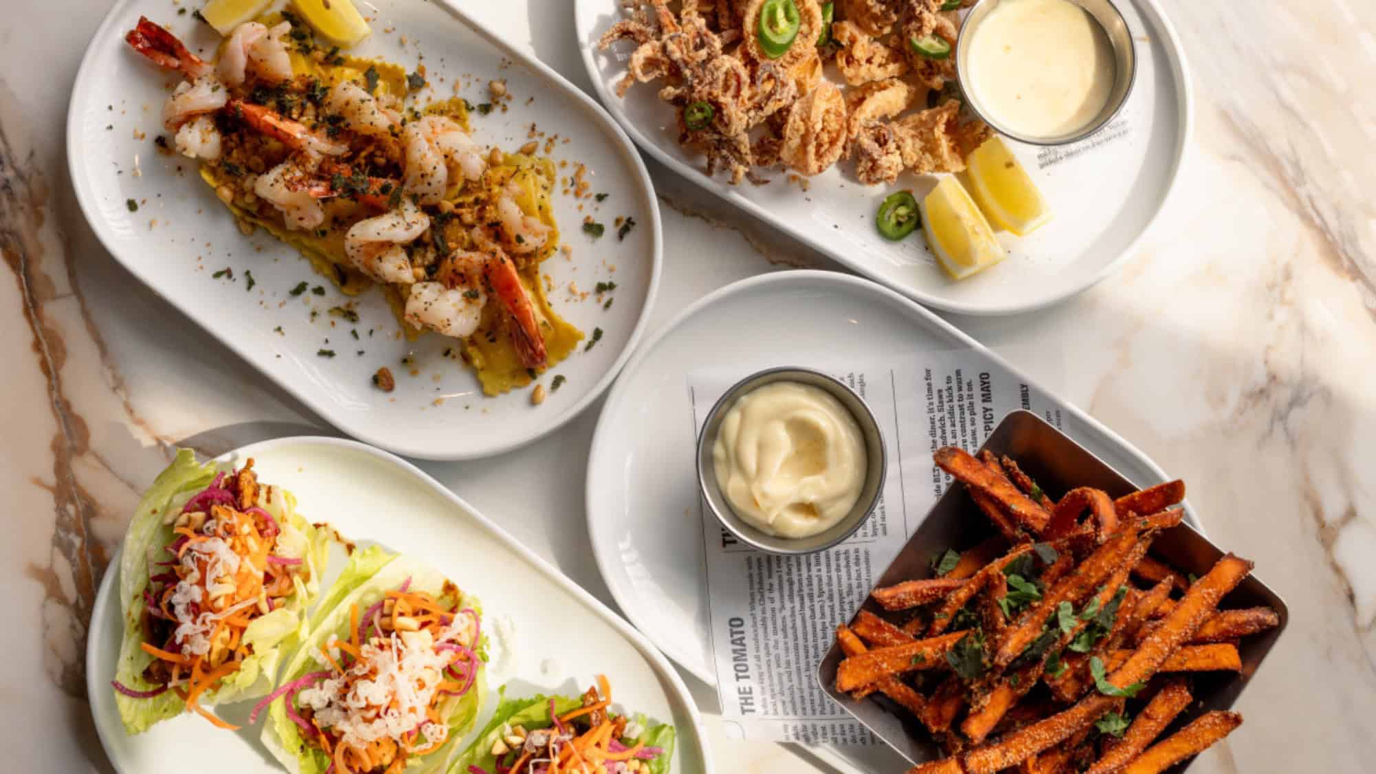 Four plates of food on a marble table: grilled shrimp, fried calamari with dipping sauce, lettuce wraps with slaw, and a basket of seasoned sweet potato fries with dip.