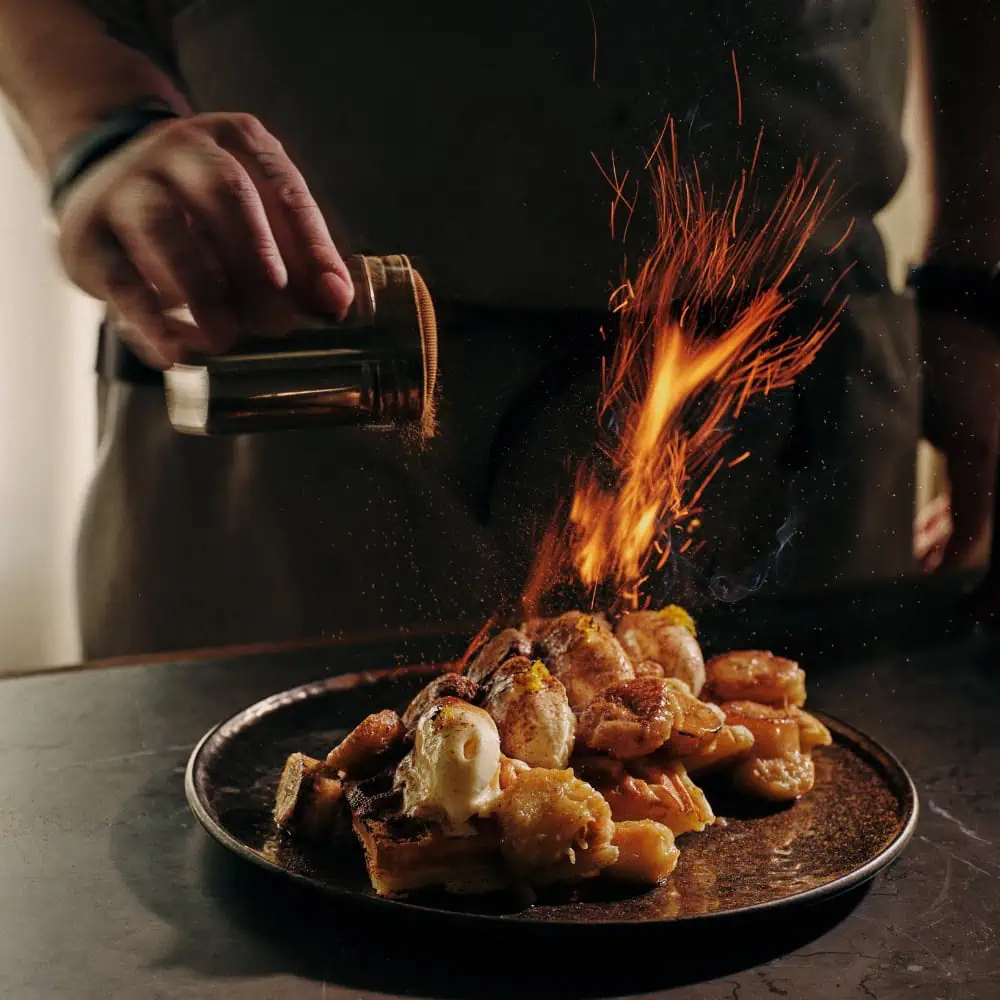 A person sprinkles spices over a plated dessert at a restaurant, causing a burst of orange sparks above the food—perfect for a festive Mother’s Day brunch.