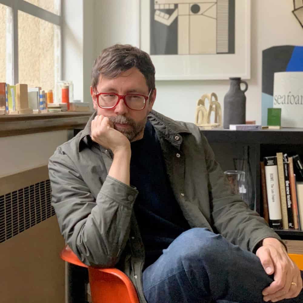 A man with glasses sits on an orange chair, resting his chin on his hand, in a room with books, art, and a window.
