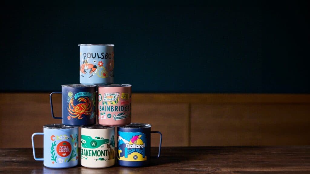Six colorful mugs with various place names and illustrations are stacked on a wooden table against a dark blue and wood-paneled background.