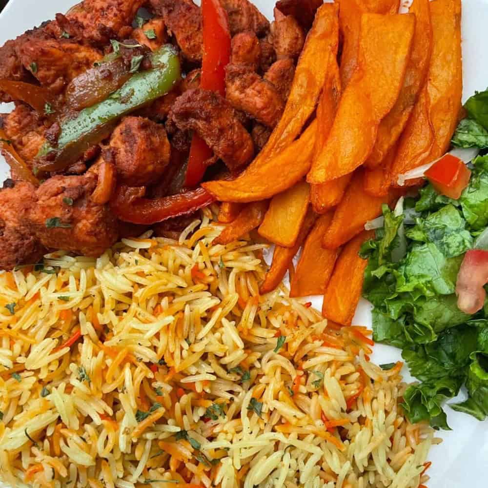 A plate with grilled chicken and peppers, sweet potato fries, seasoned rice, and a side salad with lettuce and tomato.