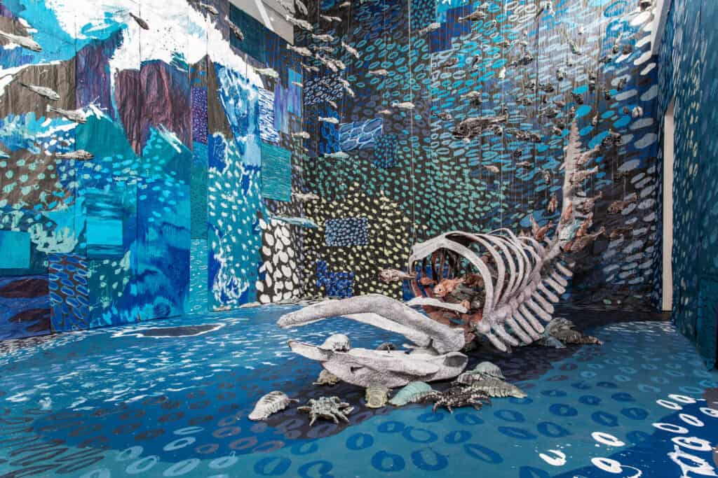 Room-sized art installation featuring a large ribcage structure, marine animal sculptures, and walls painted with abstract blue and aqua patterns resembling an underwater environment.