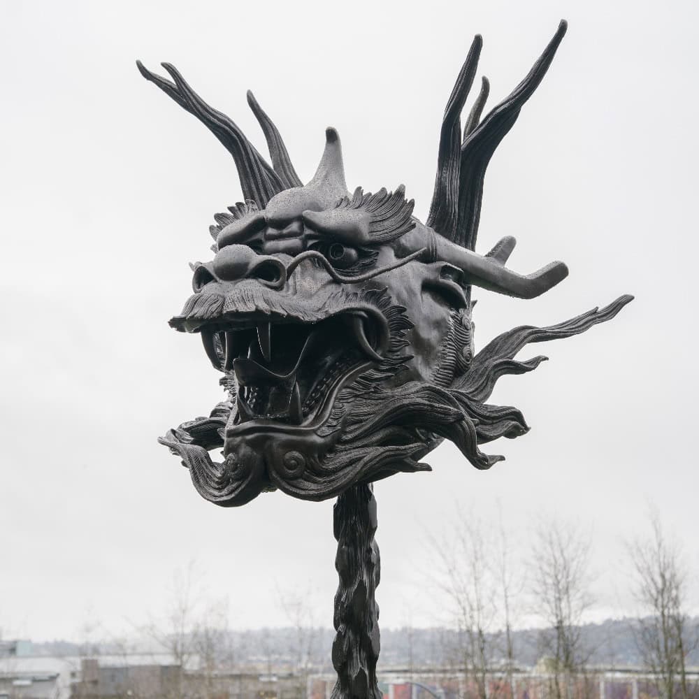 Large metal sculpture of a dragon's head with open mouth and sharp teeth, mounted on a pole outdoors, stands as a striking symbol for AANHPI Month celebrations at Seattle events, with cloudy skies and trees in the background.