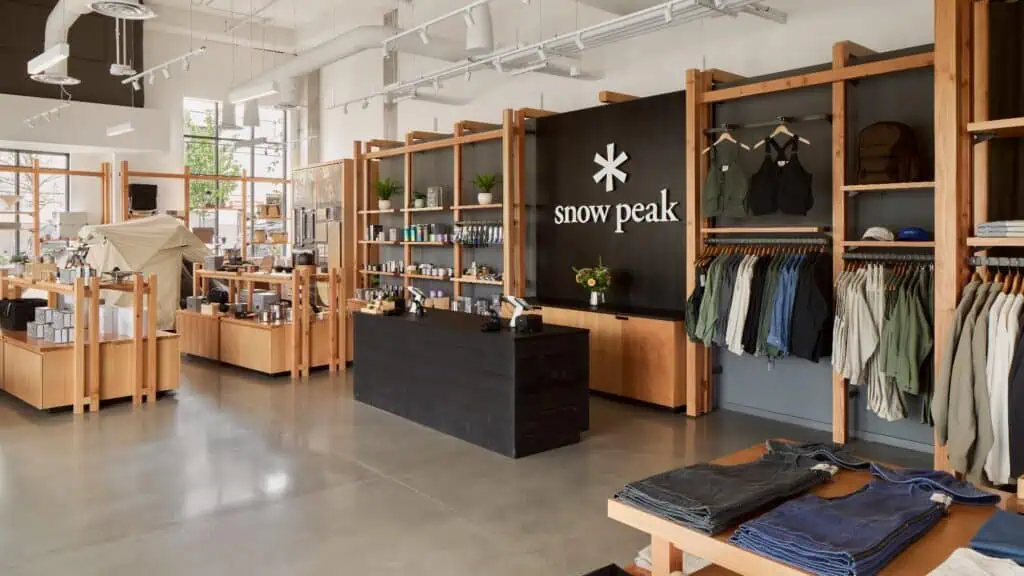 Interior of a Snow Peak retail store with wooden shelves displaying outdoor gear, clothing, and accessories; the checkout counter is centered, natural light fills the space—like a rearview mirror reflection after a Fashion Show at SIFF.