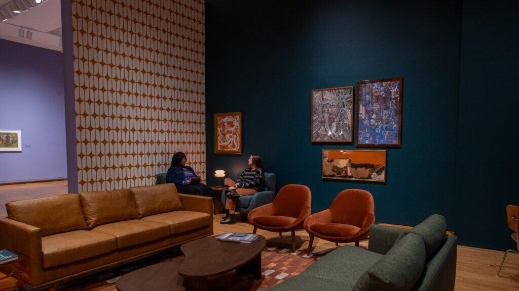 Two people sit and talk in a mid-century modern styled gallery at the Seattle Art Museum, surrounded by colorful furniture and art from Beyond Mysticism: The Modern Northwest.