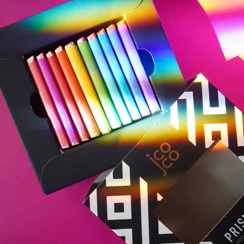 A box of jcoco chocolate bars in rainbow colors is displayed on a magenta surface, with light creating a prism effect on the packaging and wrappers.