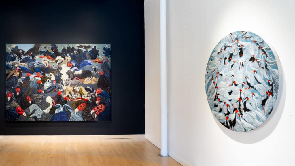 A rectangular painting of various birds hangs on a dark wall, while a circular bird-themed painting is displayed on a white wall in a gallery space with wooden floors.