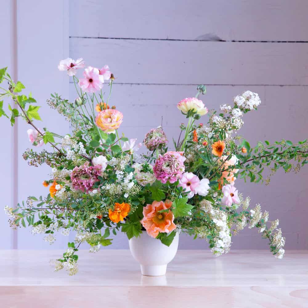 A white vase holds a loose arrangement of assorted pink, orange, and white flowers with green foliage on a light wooden table—an ideal Mother’s Day gift to brighten her special day.