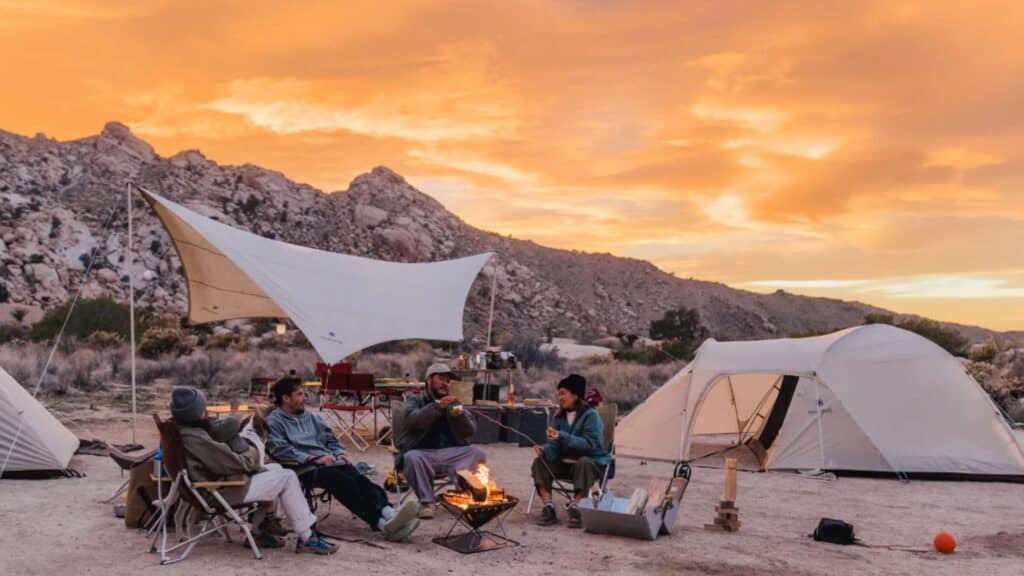 Four people sit around a campfire at a campsite with tents and canopies, surrounded by rocky hills under an orange sunset sky.