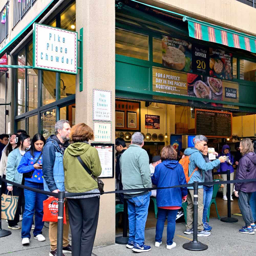People wait in line outside Pike Place Chowder, a popular eatery with a striped awning, visible signs, and menus featuring seasonal dishes and fresh spring food.
