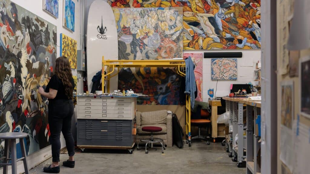 A person stands in an art studio painting on a large canvas, surrounded by colorful artwork, art supplies, and furniture.