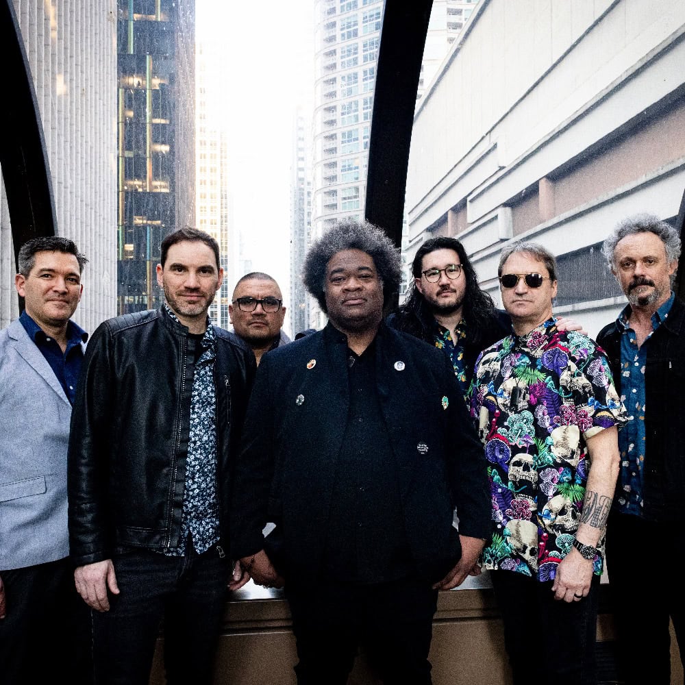 Seven men stand together in front of a cityscape, wearing various styles of clothing, including jackets, shirts, and sunglasses.