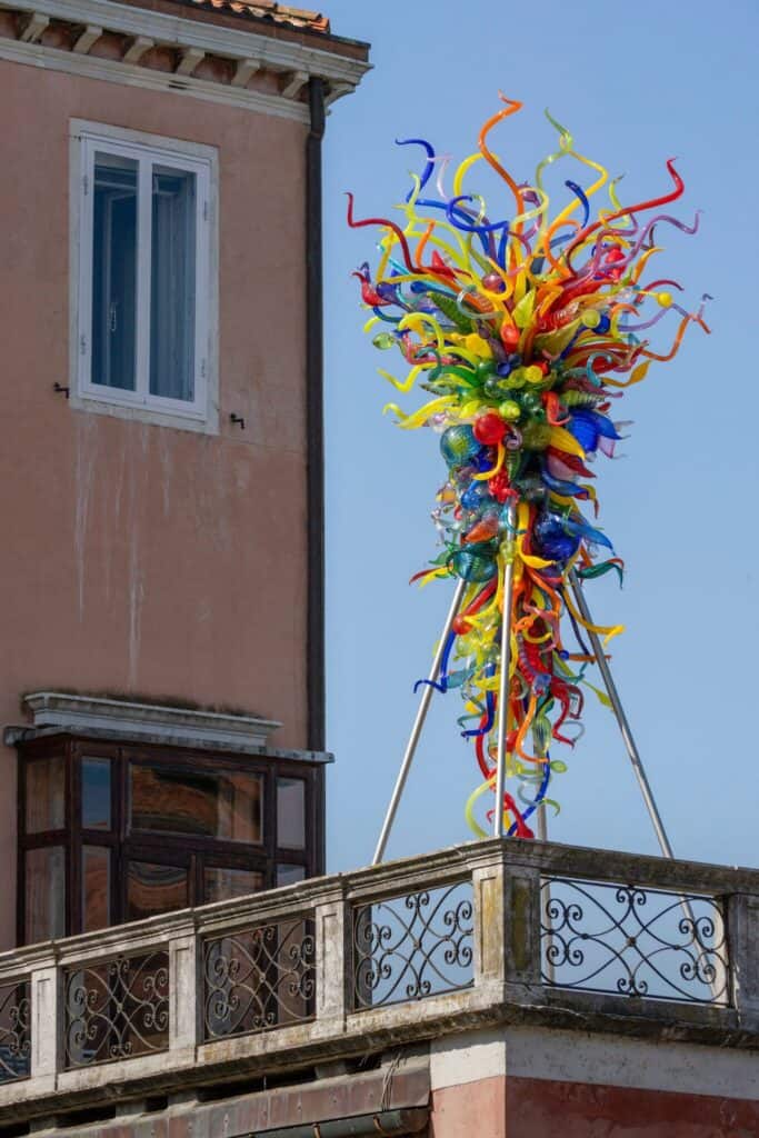 Colorful glass sculpture with twisting shapes stands on a balcony next to a peach-colored building with an ornate railing and a blue sky in the background.
