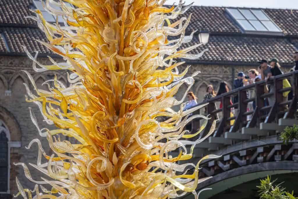 Golden glass sculpture with intricate, curling shapes stands in front of a bridge with people observing and taking photos; old brick building in the background.